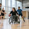 Parasportisti kopā ar treneri izpilda bumbas vingrinājumu gaišā un pieejamā sporta zālē.