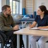 Cilvēks riteņkrēslā rehabilitācijas kabinetā pārrunā individuālo plānu ar speciālisti