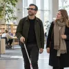Vīrietis ar balto spieķi un viedajām brillēm kopā ar pavadoni pārvietojas gaišā bibliotēkā