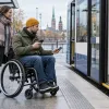 Vīrietis riteņkrēslā ar transporta karti un tālruni gatavojas iekāpt zemās grīdas sabiedriskajā transportā.
