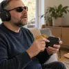 Neredzīgs vīrietis ar austiņām lieto kompaktu audio atskaņotāju ar izteikti taustāmām pogām.