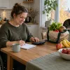 Sieviete riteņkrēslā pie virtuves galda plāno pārtikas pirkumus līdzās iepirkumu somai ar produktiem.