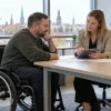Vīrietis riteņkrēslā kopā ar sociālo darbinieci pie pieejama galda pārrunā plānu planšetē.