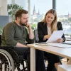 Vīrietis riteņkrēslā un karjeras konsultante pie galda kopīgi pārskata darba pieteikuma dokumentus